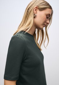 Mladá žena s dlouhými blond vlasy má na sobě tmavě zelené tričko s krátkým rukávem a stříbrné kruhové náušnice, dívá se dolů proti jednoduchému světlému pozadí.