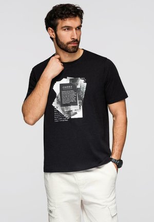 Mann mit Bart, der ein schwarzes T-Shirt mit Grafik und weiße Hose trägt, seitlich schaut, eine Hand nahe am Hals und die andere in der Tasche hat.