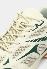 Λευκό αθλητικό παπούτσι με πράσινες λεπτομέρειες, διαθέτει επάνω μέρος από δίχτυ για αερισμό, υφασμένα κορδόνια και ελαστική σόλα για κράτημα.