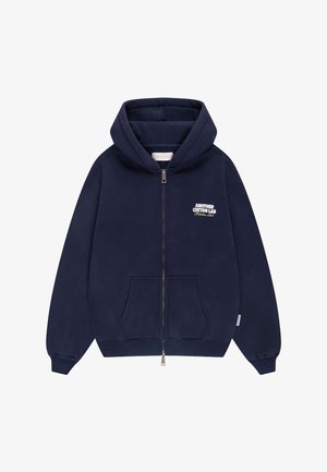 Sweat à capuche bleu marine zippé avec poches avant et texte blanc "Another Cotton Lab Pilates Lab" sur le côté gauche de la poitrine.