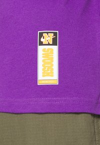 Lila bomulls t-shirt med en rektangulär etikett med "SWOOSH" i gult och "N"-logotyp ovanför. Skjortan har en jämn textur och snygg sömnad.