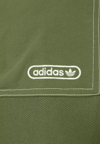 Olivgrön tygpatch med en broderad vit Adidas-logotyp med trefoil-design, kantad av vit sömnad.