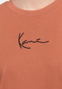 Camiseta de color óxido hecha de tela suave, con texto bordado en negro que dice "Kani" cerca del cuello. Diseño sencillo de cuello redondo.
