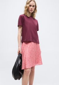 Γυναίκα που φοράει μπορντό τοπ με κοντά μανίκια και φούστα με ροζ λουλούδια, κρατώντας μια μαύρη τσάντα, μπροστά σε ένα απλό φωτεινό φόντο.