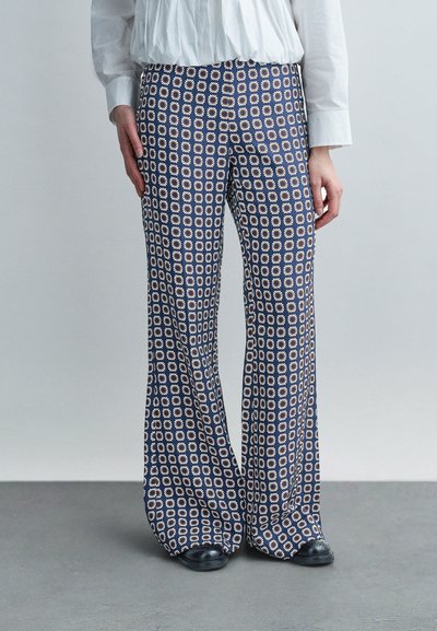 Femme portant un pantalon à jambes larges avec un motif géométrique de marguerites bleu et blanc, associé à un chemisier blanc et des chaussures noires.