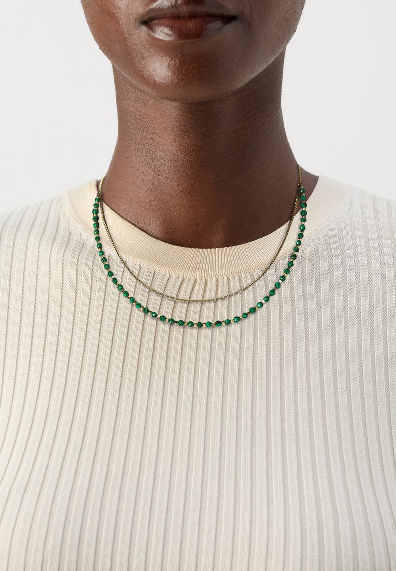 Guld-tone halskæde lagdelt med grønne perle detaljer, båret over en cremefarvet ribbet sweater. Nærbillede af nakkeområdet der fremhæver smykker og stof tekstur.