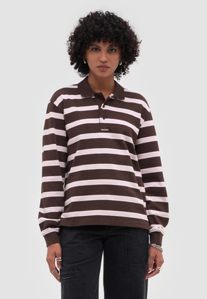 Osoba s kudrnatými vlasy, která má na sobě pruhované dlouhé tričko s límečkem v hnědé a světle růžové barvě a černé kalhoty, stojí před jednoduchým pozadím.