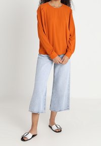 Orange langærmet bluse med plisseret skulderdetalje, parret med lys blå brede denimjeans og sort-hvide slidesandaler.