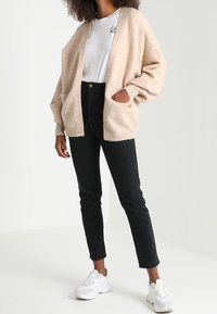 Krémový oversized pletený kardigan s otevřeným předním dílem, dvěma kapsami a žebrovanými manžetami, nošený přes bílé tričko a černé skinny džíny. Bílé tenisky.