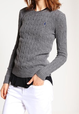 Nainen, jolla on päällään istuva harmaa kaapelineulottu neulepaita, jossa on pieni sininen logo, valkoiset housut ja musta kerrospaita alla.