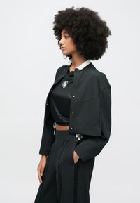 Μαύρο κοντό σακάκι με κουμπιά-πατιλέτα, με λευκό γιακά. Φορεμένο πάνω από μαύρο κοντομάνικο πουκάμισο με υφή. Μαύρο παντελόνι.