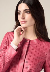 Giacca in pelle perforata rosa con collo rotondo, chiusura a pressione discreta e polsini a coste. Tessuto liscio e fori uniformemente distribuiti.