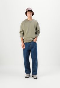 Sudadera verde oliva con puños acanalados, combinada con jeans azules holgados. La persona lleva un sombrero de cubo morado claro y zapatillas grises.