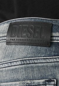 Svart läderetikett på blekt blå denim, präglad med "DIESEL" och "FÖR FRAMGÅNGSRIKT LIV." Synlig sömnad längs midjebandet.