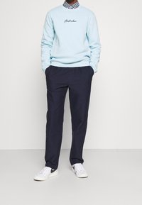 Sudadera azul claro con bordado de "Ámsterdam", combinada con pantalones oscuros y zapatillas blancas. Presenta un cuello a cuadros por debajo.