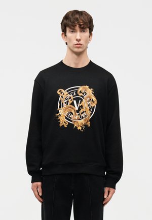 Νέος άνδρας με κοντά καστανά μαλλιά, φορώντας μαύρο φούτερ Versace Jeans Couture με χρυσά μπαρόκ μοτίβα και λογότυπο, στέκεται μπροστά σε λευκό φόντο.