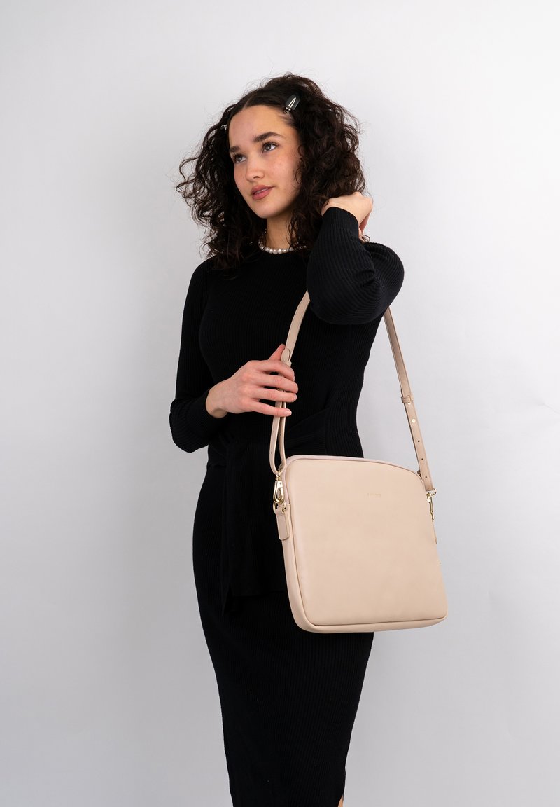 Beige læder skuldertaske med firkantet form og justerbar rem. Har guldhardware og minimalistisk design, holdt af en person i en sort kjole.