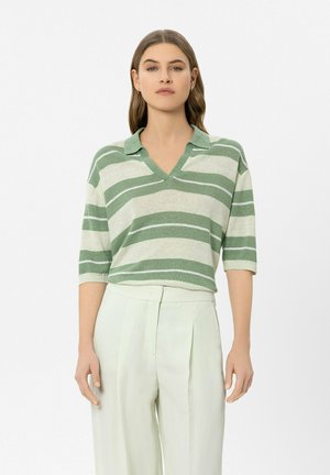 Woman wearing a green and white striped collared knit top with elbow-length sleeves and high-waisted pale trousers against a white background.