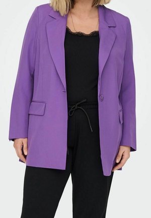 Frau trägt einen violetten Ein-Knopf-Blazer über einem schwarzen Top mit Spitzenbesatz und schwarzen Hosen mit Kordelzug, steht vor weißem Hintergrund.