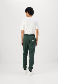 Dark green jogger pants with elastic cuffs, drawstring waist, and a small label on the back pocket, paired with a white short-sleeve shirt.