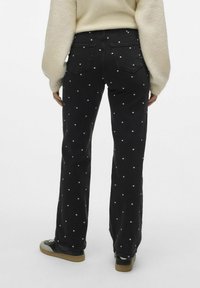 Sorte jeans med hvite prikker, utsvingt design, middels høyde i midjen, to baklommer, brukt med en kremfarget genser og svarte joggesko.