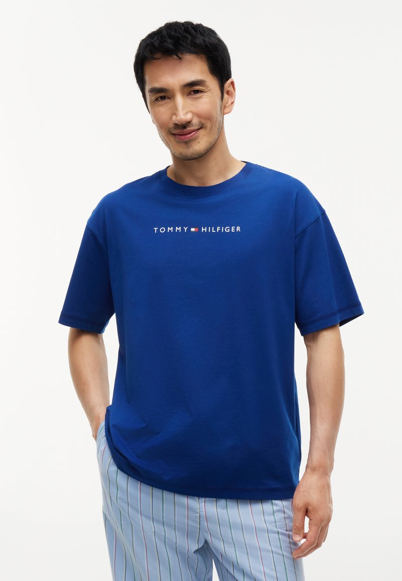 Χαμογελαστός άνδρας που φοράει μπλε T-shirt Tommy Hilfiger και ανοιχτό μπλε ριγέ παντελόνια με το ένα χέρι στην τσέπη του σε απλό φόντο.