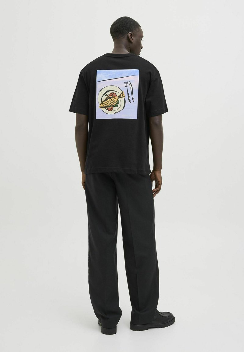 Sort bomuld t-shirt med et farverigt grafik af en tallerken med fisk og bestik på ryggen. Matcher med løse sorte bukser og sko.