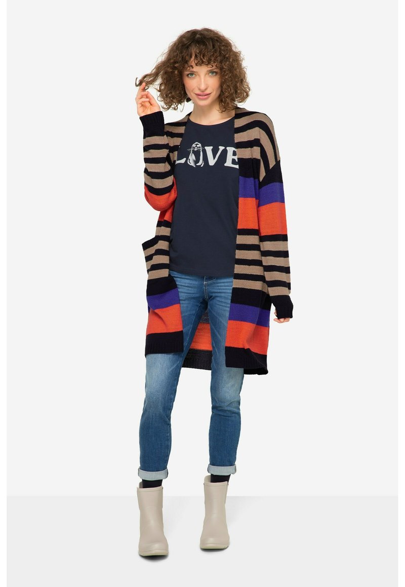 Färgglad lång cardigan med horisontella ränder i orange, blått, svart och beige, bärs över en marinblå t-shirt, ihop med jeans och beige stövlar.