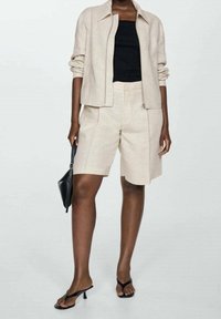 Beige linned blazer med en afslappet pasform, parret med matchende beige skræddersyede shorts. Sort tanktop under. Sort firkantet håndtaske.