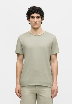 Νέος άνδρας με κοντά σγουρά σκούρα μαλλιά που φοράει ανοιχτό πράσινο κοντομάνικο μπλουζάκι και αντίστοιχο παντελόνι, στέκεται μπροστά σε απλό λευκό φόντο.