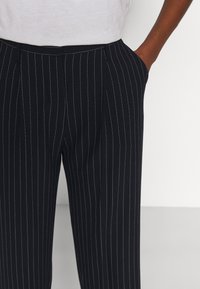 Námořnické modré kalhoty s pruhy a úzkým střihem, které mají boční kapsy a hladkou texturu. Pasová guma leží mírně nad boky.