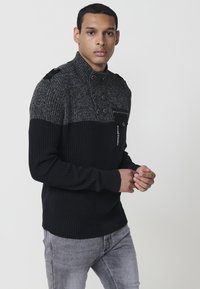 Suéter negro acanalado con cuello alto; cuenta con un hombro texturizado gris, tapeta de dos botones y un bolsillo con cremallera en el pecho.