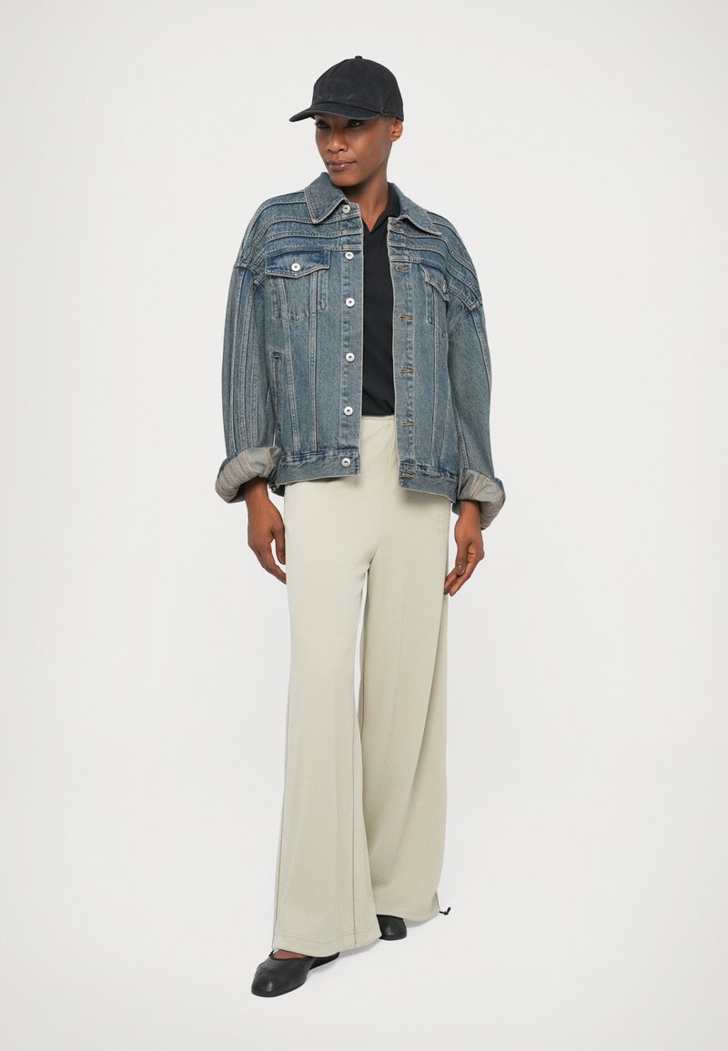 Veste en denim avec un lavage bleu clair, deux poches avant et des boutons métalliques, associée à un pantalon large crème et des accessoires noirs, y compris une casquette et des chaussures.