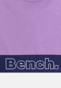 Fialové bavlnené tričko s rebrovanou textúrou a okrúhlym výstrihom. Na spodnej strane je tmavomodrý pruh s bielym vyšitým logom "Bench.".