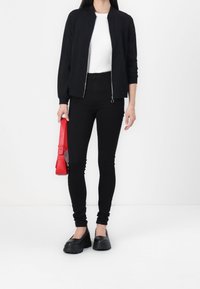 Persona che indossa una giacca nera con zip, una maglietta bianca, pantaloni neri e scarpe chunky nere, mentre tiene una borsa rossa su uno sfondo bianco.