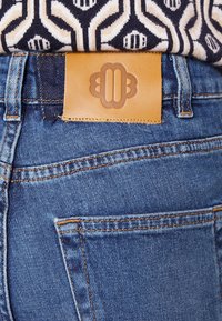 Farkut denim, joissa on vaalea nahkapatentti, jossa on kohokuvioitu kuvio vyötäröllä. Huomiota herättävä oranssi ompelu ja klassinen viiden taskun tyyli.