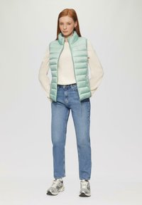 Hellgrüner Puffervest mit horizontalen Nähten und hohem Kragen, über einem cremefarbenen Pullover, kombiniert mit blauen Jeans und silbernen Sneakers.