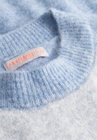 Lichtblauwe wollen trui met een geribde kraag, voorzien van een label in het roze met de merknaam. De stof lijkt zacht en licht donzig.