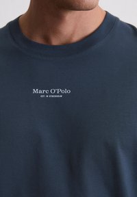 Άνδρας που φοράει μπλε κωδικό μπλουζάκι με λευκό λογότυπο Marc O’Polo και κείμενο "Est. στη Στοκχόλμη" στο στήθος.