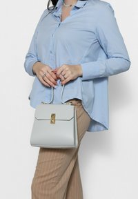 Borsa a tracolla grigia chiara con design strutturato, chiusura dorata e due manici superiori, tenuta da un modello che indossa una camicia azzurra chiara e pantaloni beige a righe.