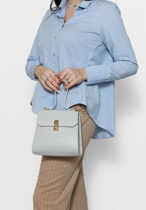 Sac à main gris clair au design structuré, avec un fermoir doré et des poignées supérieures doubles, porté par un mannequin dans une chemise bleu clair et un pantalon beige à rayures.
