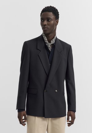 Mann trägt schwarzen zweireihigen Blazer, marineblaues Hemd, beige Hose und gestreiften Halstuch, steht vor schlichtem grauem Hintergrund.