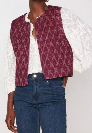 Mujer con blusa blanca de encaje con mangas abullonadas, chaleco sin mangas burdeos con estampado y vaqueros azules de talle alto, mano en el bolsillo.