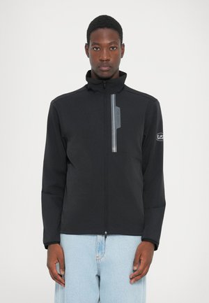 Schwarze Jacke mit hohem Kragen, Reißverschluss vorne, reflektierendem Streifen und Logodetails auf der Brust und dem Ärmel. Glattes Material, taillierter Schnitt.