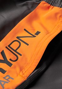 Zwarte sportshorts met een oranje paneel met gedurfde zwarte tekst. De stof heeft een gladde textuur en de elastische tailleband is zichtbaar.