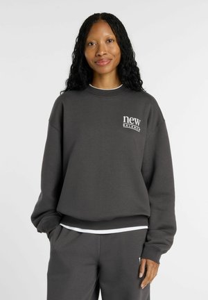 Mujer con cabello largo y rizado que lleva una sudadera New Balance gris oscuro y pantalones a juego, de pie con una mano en el bolsillo.