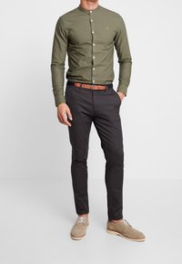 Mężczyzna ubrany w oliwkowozieloną koszulę na guziki, ciemnoszare spodnie, brązowy pasek i beżowe zamszowe buty, stojący z jedną ręką w kieszeni.