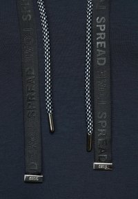 Marinblå huvtröja med dubbla snören; ett tjockt svart med präglad text och ett tunt rutigt. Silverhängslen i ändarna. Mjuk textur.