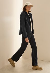 Svart huva jacka, krämfärgad topp, mörka jeans och beige skor. Jackan har en dragkedja och två fickor, med synlig sömnad på jeansen.