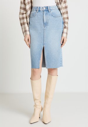 Femme portant une jupe en jean bleu clair avec une fente frontale, des bottes beige montant jusqu'aux genoux, un haut blanc côtelé et une chemise à carreaux.